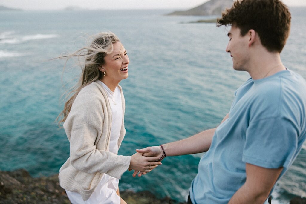 A woman smiles and laughs at a man as they hold hands on Makapuu Lookout in Hawaii before their surprise proposal