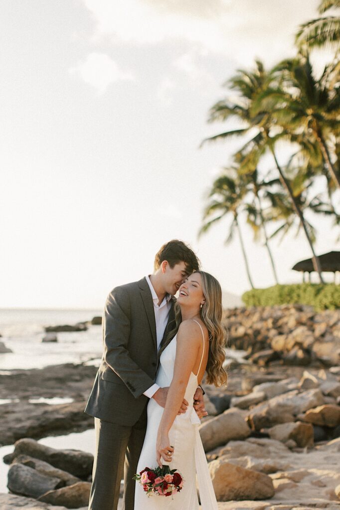 A couple on a beach in Hawaii embrace and laugh, the woman is holding a bridal bouquet and is in her wedding dress
