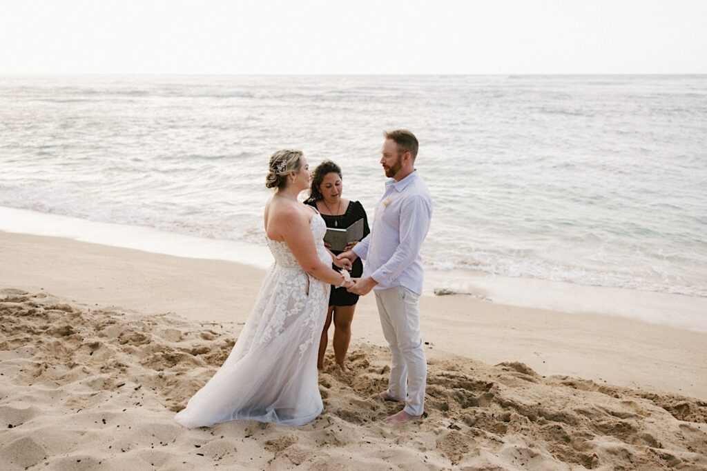 A bride and groom hold hands as their officiant speaks during their elopement on a beach in Hawaii