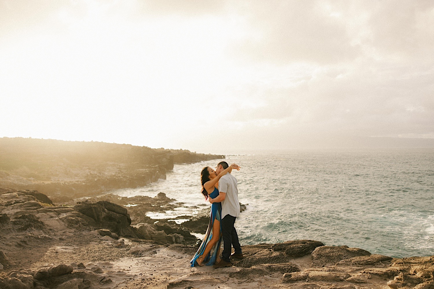 During their engagement session in Hawaii a couple kiss and embrace while on a rocky cliff that looks out over the ocean during sunset