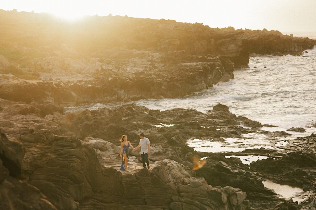 During their engagement session in Hawaii a couple hold hands and walk while on a rocky cliff that looks out over the ocean during sunset