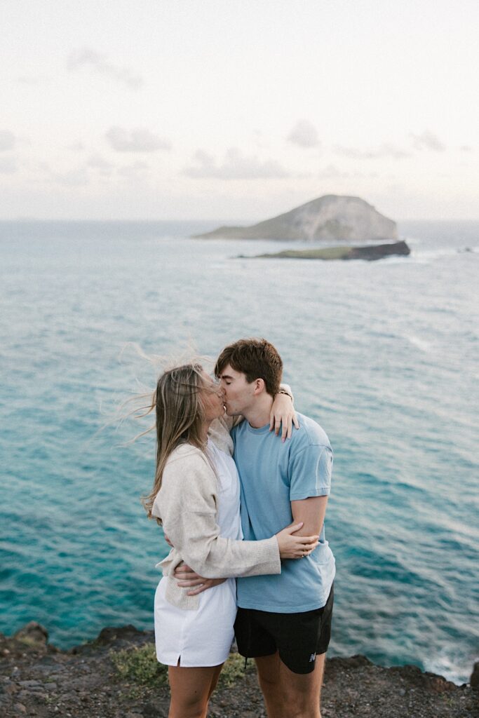 A now engaged couple kiss one another on a cliff in Hawaii looking out over the ocean and the islands