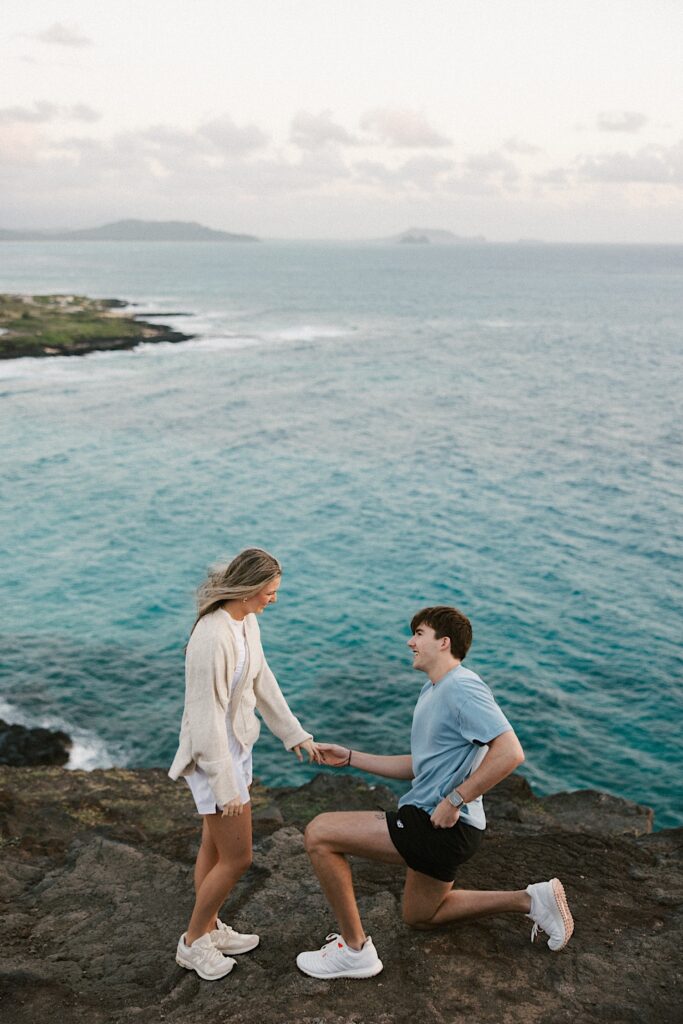 A man gets down on one knee and pulls something out of his pocket while holding a woman's hand to propose to her on a cliff in Hawaii