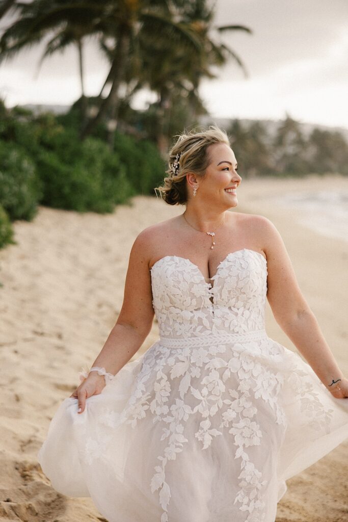 A woman in a wedding dress smiles as she walks down a beach and looks to the right at the ocean