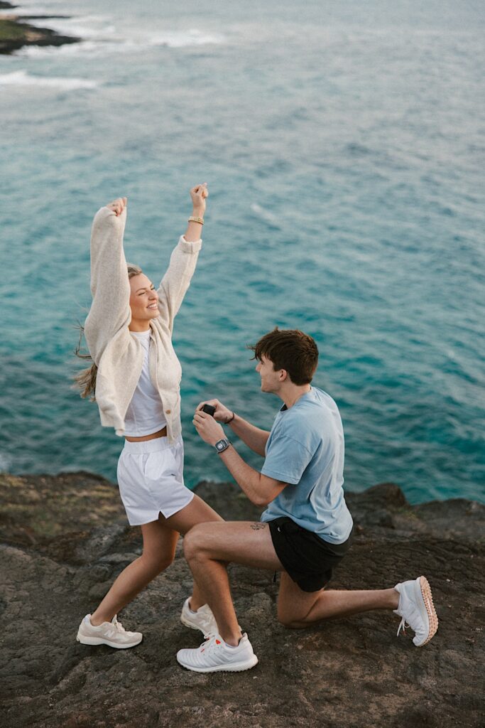 A woman smiles and throws her arms in the air as a man in front of her proposes to her atop a cliff in Hawaii looking out over the ocean