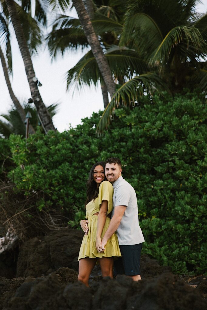 A couple smile at the camera as they embrace and hold each others hand with palm trees and other greenery behind them.