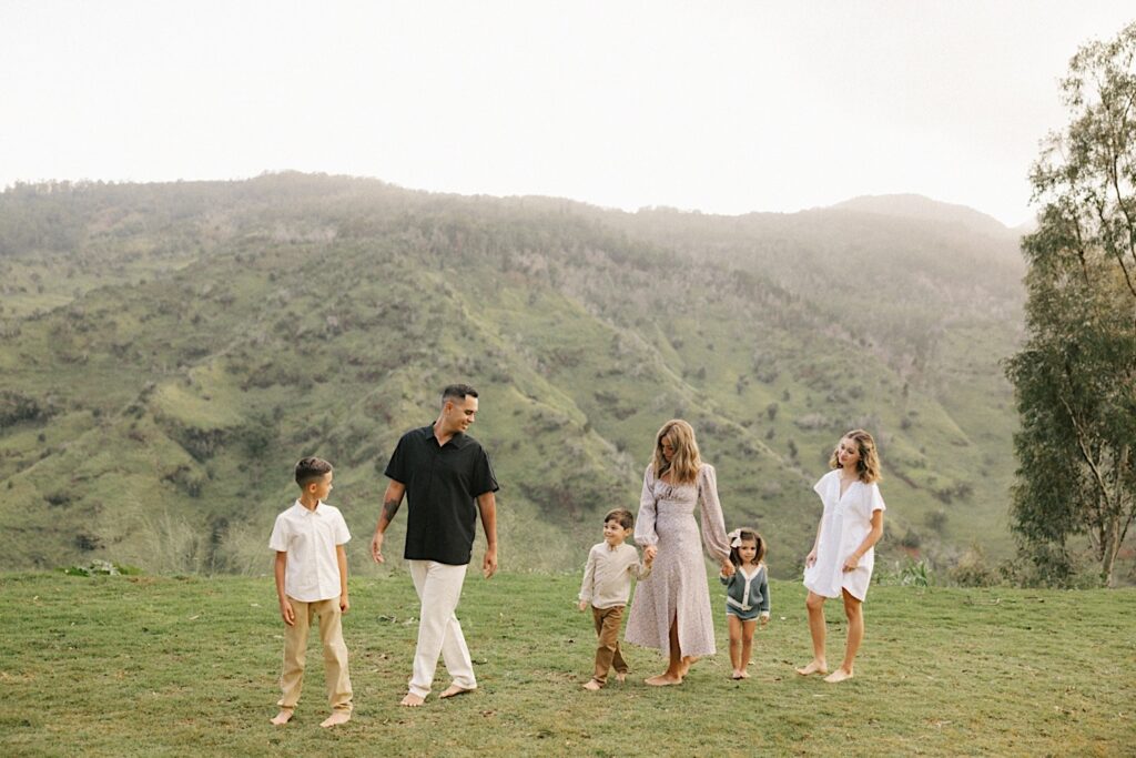 A couple walks with their four children during their family session on Oahu, the youngest two are holding their mother's hands, in the background is a lush green mountain