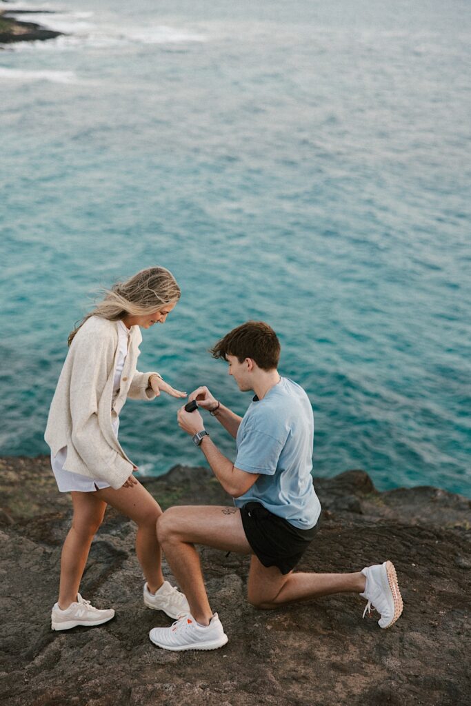 A man puts a ring on his now fiancée after his proposal to her on a cliff in Hawaii looking out over the ocean 