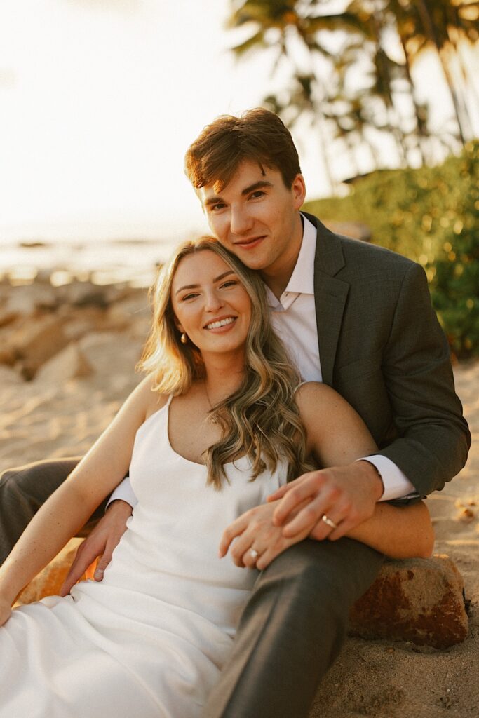 A couple sit on a beach in their wedding attire and smile at the camera with palm trees and a sunset behind them