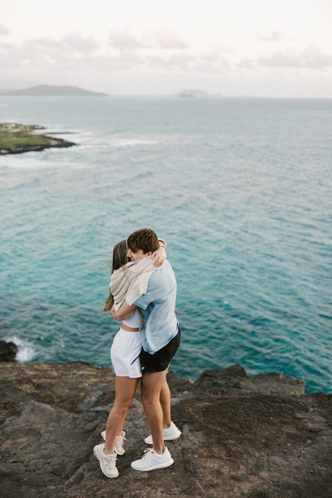 A now engaged couple hug one another on a cliff in Hawaii looking out over the ocean and the islands