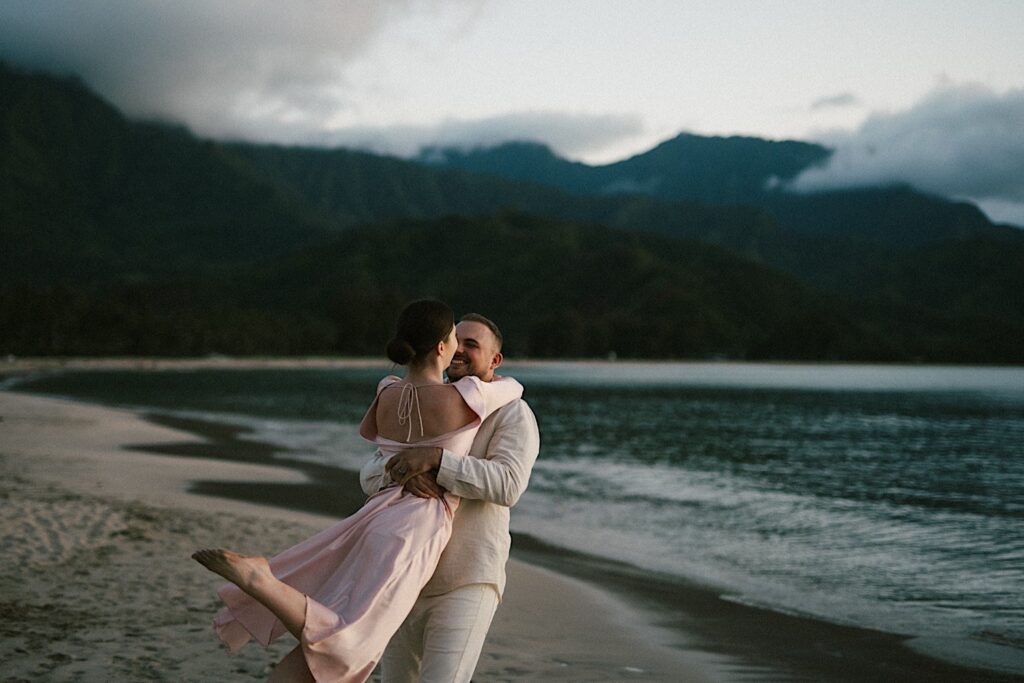 A man smiles as he picks up and swings his fiancée around while on the beach during their engagement session in Hawaii