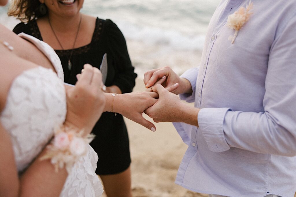 Close up photo of a groom putting a wedding ring on the bride as the officiant smiles in the background during their beach elopement in Hawaii