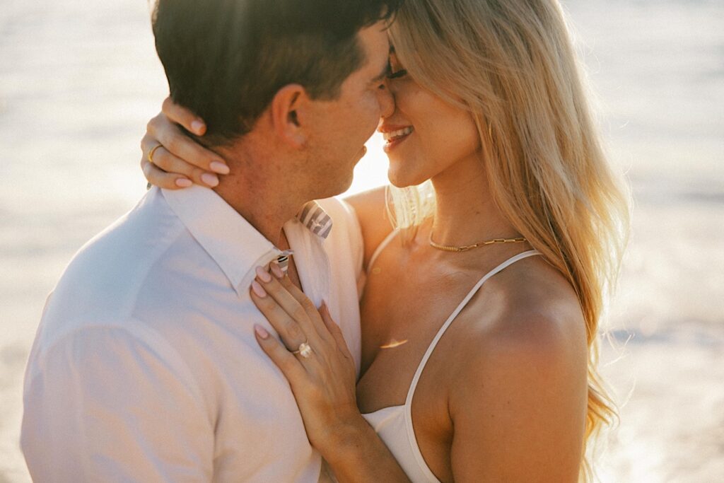 During their engagement session a  couple embrace and are about to kiss while the sun sets on a beach in Hawaii