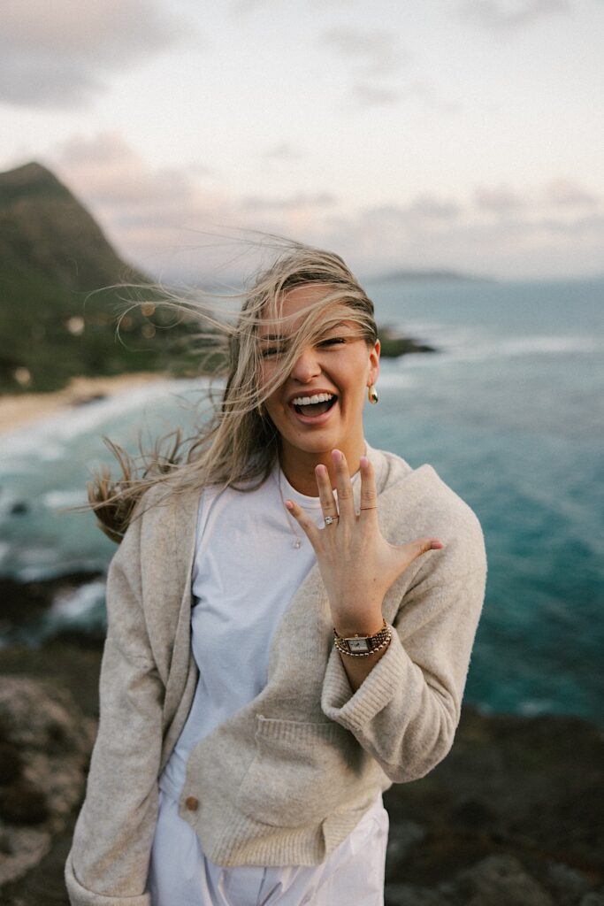 A woman on a cliff in Hawaii looking out over the ocean and the island smiles as she shows off her engagement ring to the camera