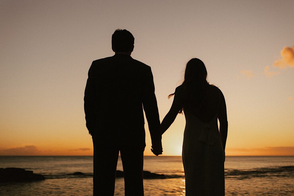A silhouette of a man and a woman holding hands while looking out over the ocean as the sun sets