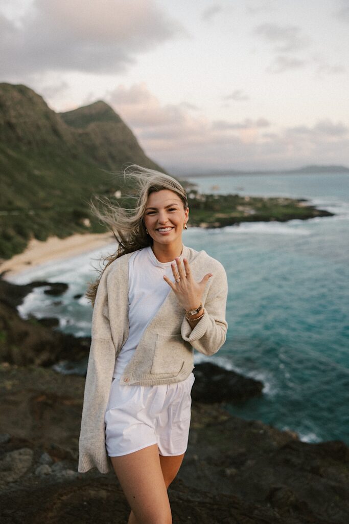 A woman on a cliff in Hawaii looking out over the ocean and the island smiles as she shows off her engagement ring to the camera