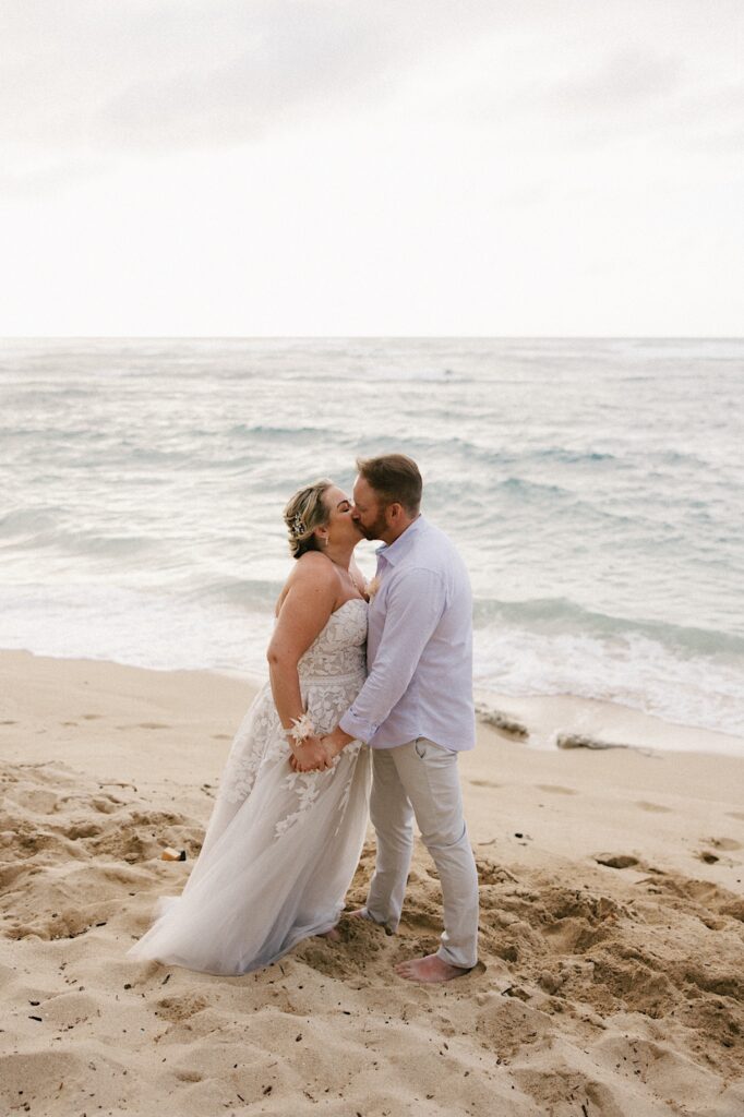 A bride and groom kiss one another while standing on the beach of Hawaii as the ocean waves crash behind them