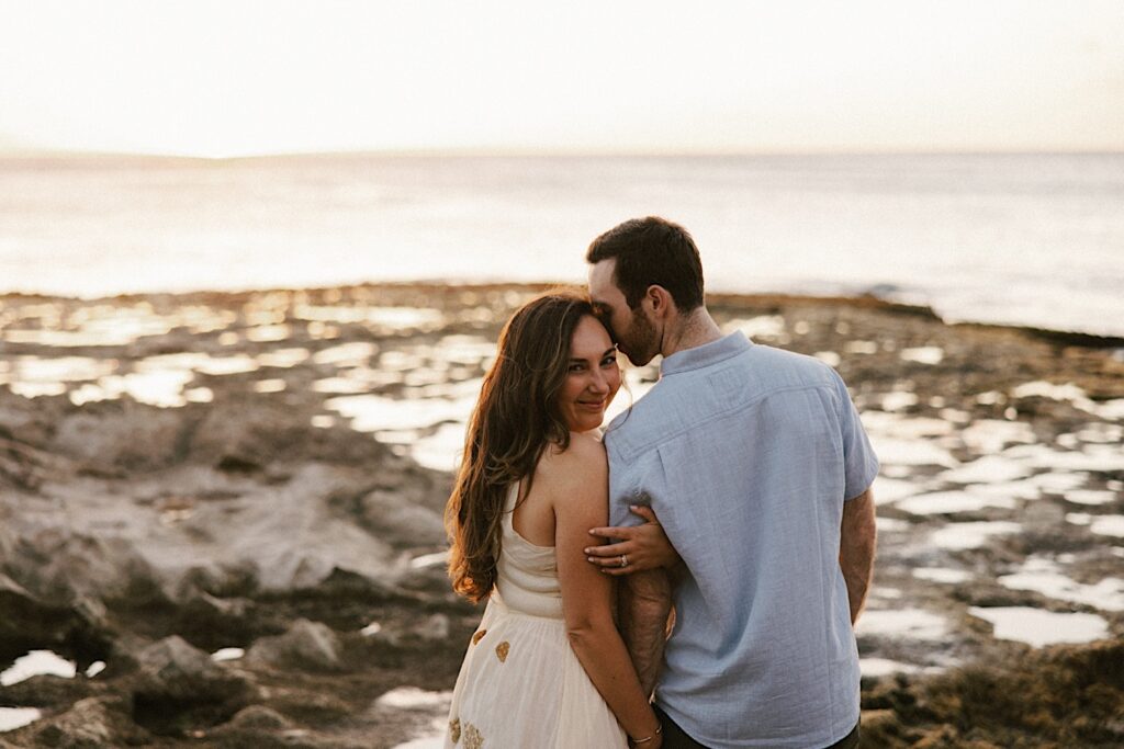 A woman smiles at the camera while hugging a mans arm as he kisses her during their engagement session in Hawaii, in the background is the ocean