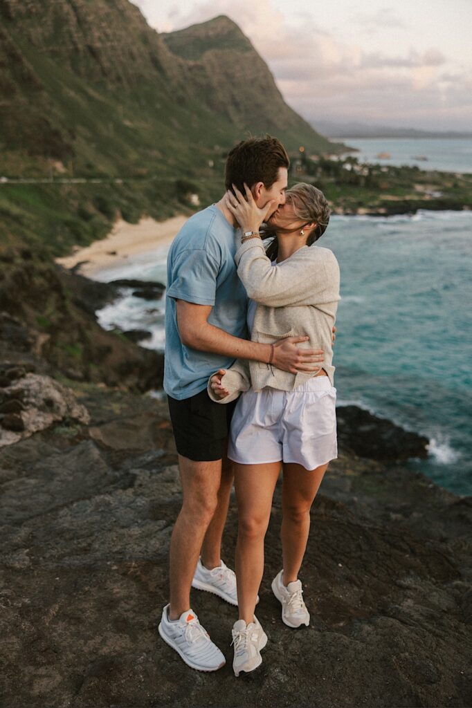 A couple on a cliff in Hawaii that looks out over the ocean and the landscape of the island kiss one another after their proposal