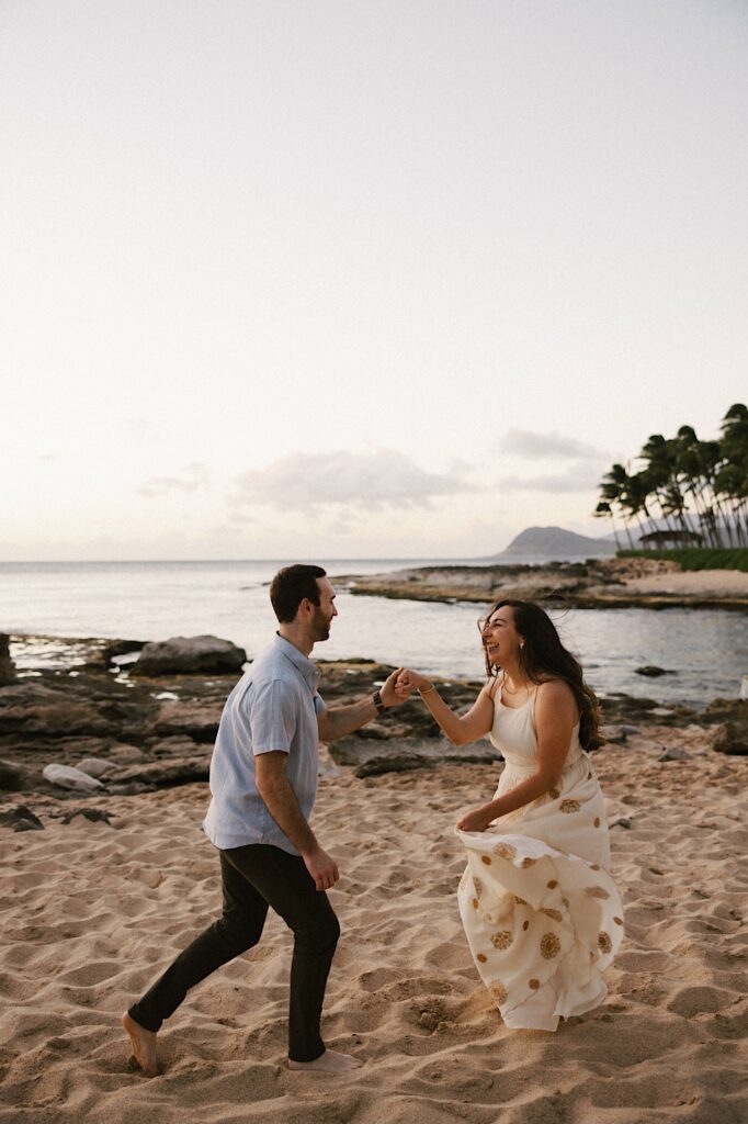 A man and woman dance in the sand while holding hands with the Hawaiian islands in the background