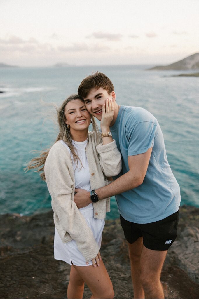 A couple embrace and smile at the camera while standing on a cliff in Hawaii that looks out over the ocean