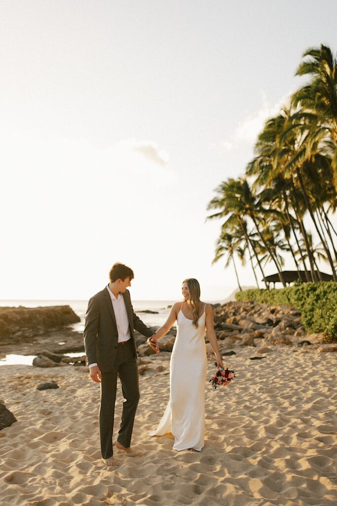 A bride and groom walk hand in hand down a beach in Hawaii and smile at one another as the sun sets