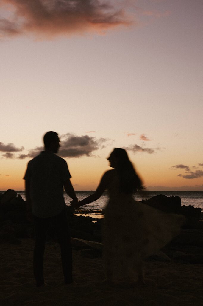 The silhouette of a man and a woman holding hands during a sunset on Hawaii with the ocean in the background