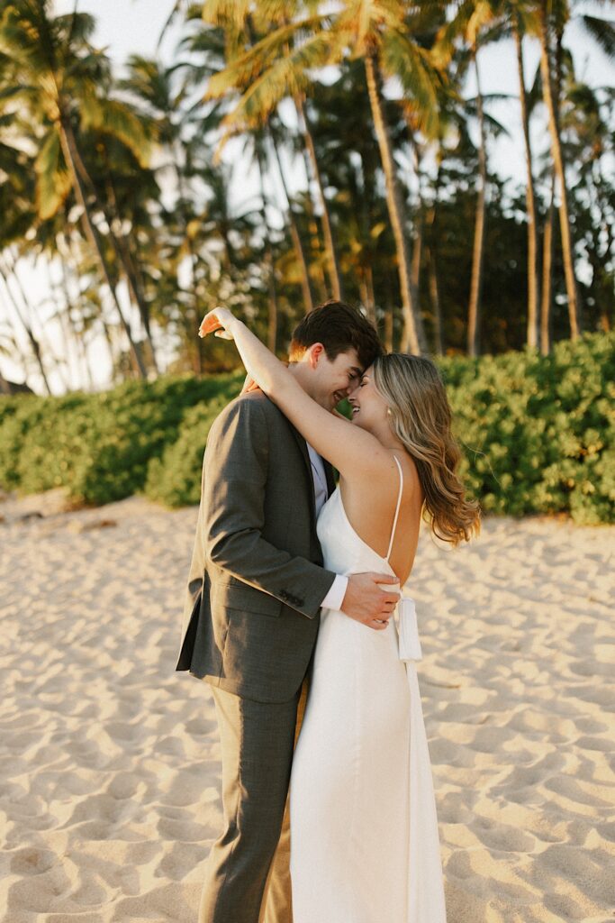 A bride and groom embrace and are about to kiss while standing on a beach in Hawaii with palm trees behind them