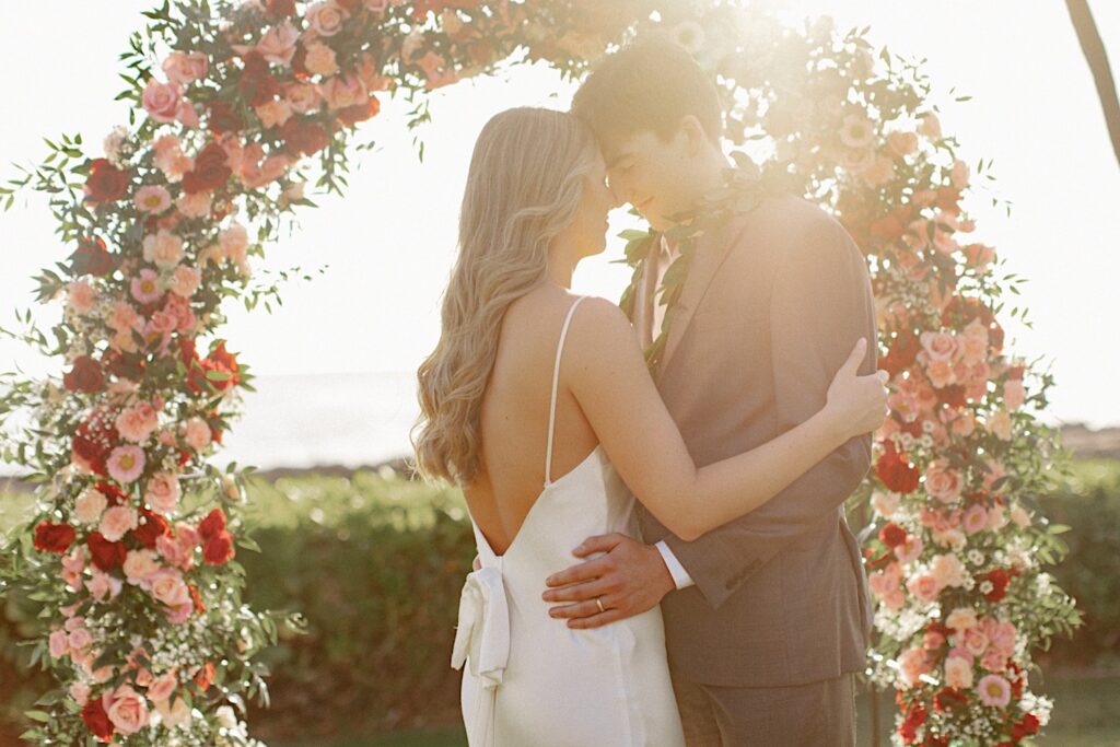 A bride and groom embrace underneath an arch of flowers while the sun sets over the ocean behind them