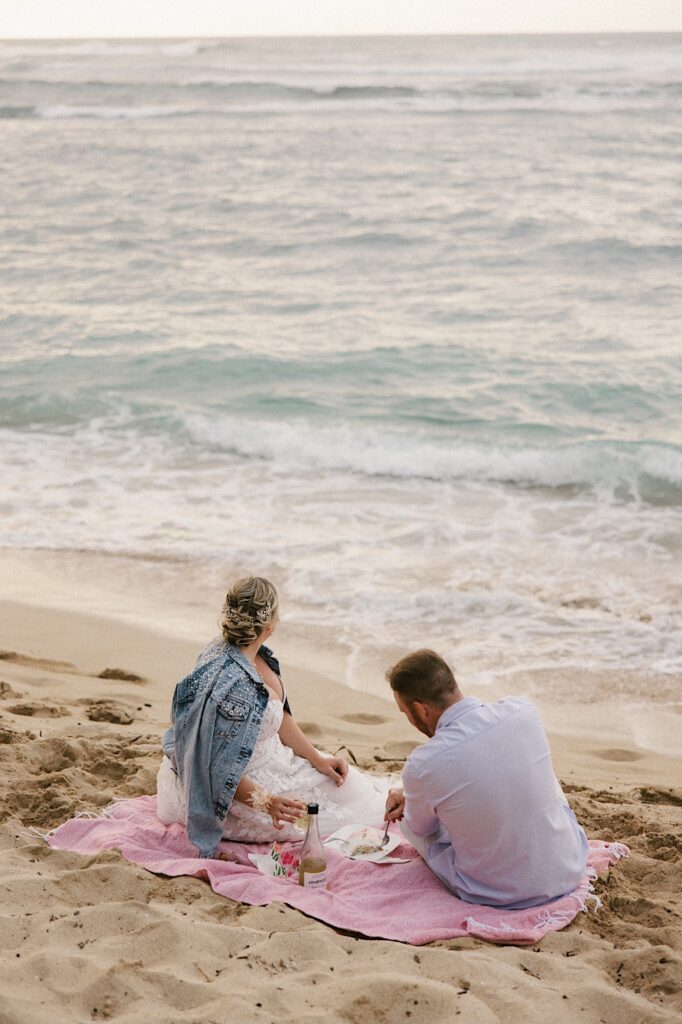A bride and groom sit on a pink towel on a beach while having a picnic looking out over the ocean of Hawaii