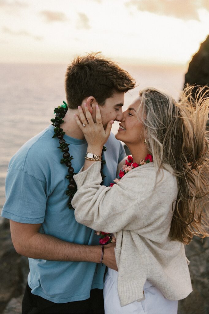 A couple on a cliff in Hawaii that looks out over the ocean and the landscape of the island lean in to kiss one another after their proposal