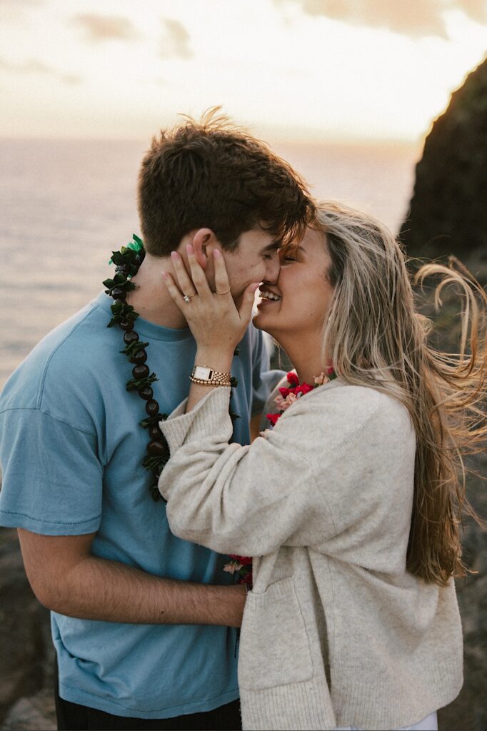 A couple on a cliff in Hawaii that looks out over the ocean and the landscape of the island lean in to kiss one another after their proposal
