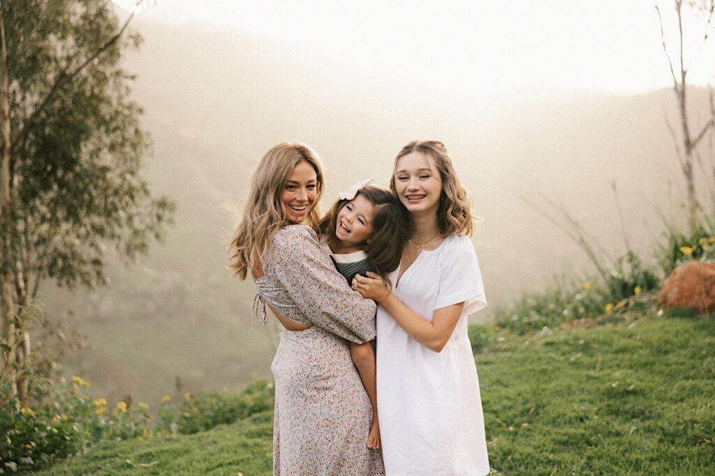 During a family session a mother and her two daughters laugh at the camera, the youngest is being held by the mother, in the background are the foggy mountains of Oahu