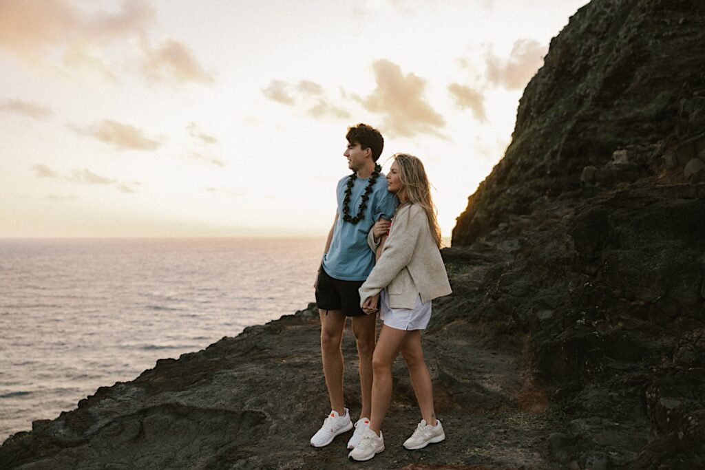 After their proposal on Makapuu Lookout in Hawaii a couple stand holding hands together and look out over the ocean as the sun sets