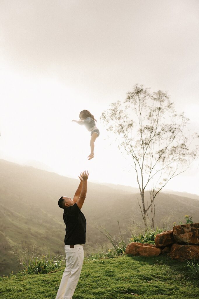 A father throws his daughter up in the air and extends his arms up to catch her, in the background are the foggy mountains of Oahu
