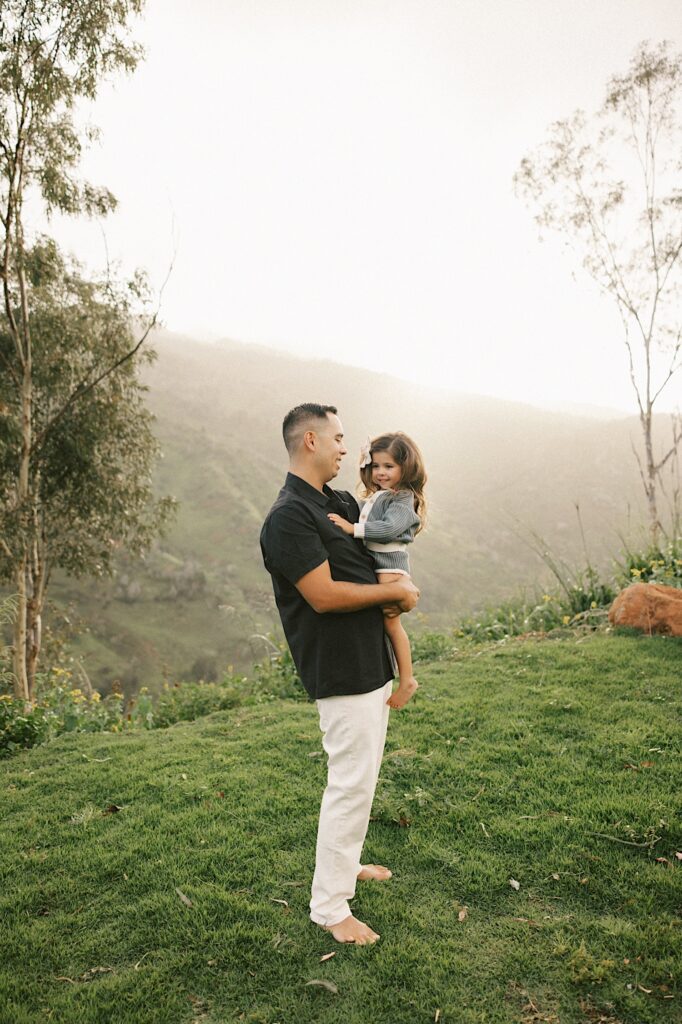 A father holds his youngest daughter in his arms and smiles at her with the foggy mountains of Oahu behind them