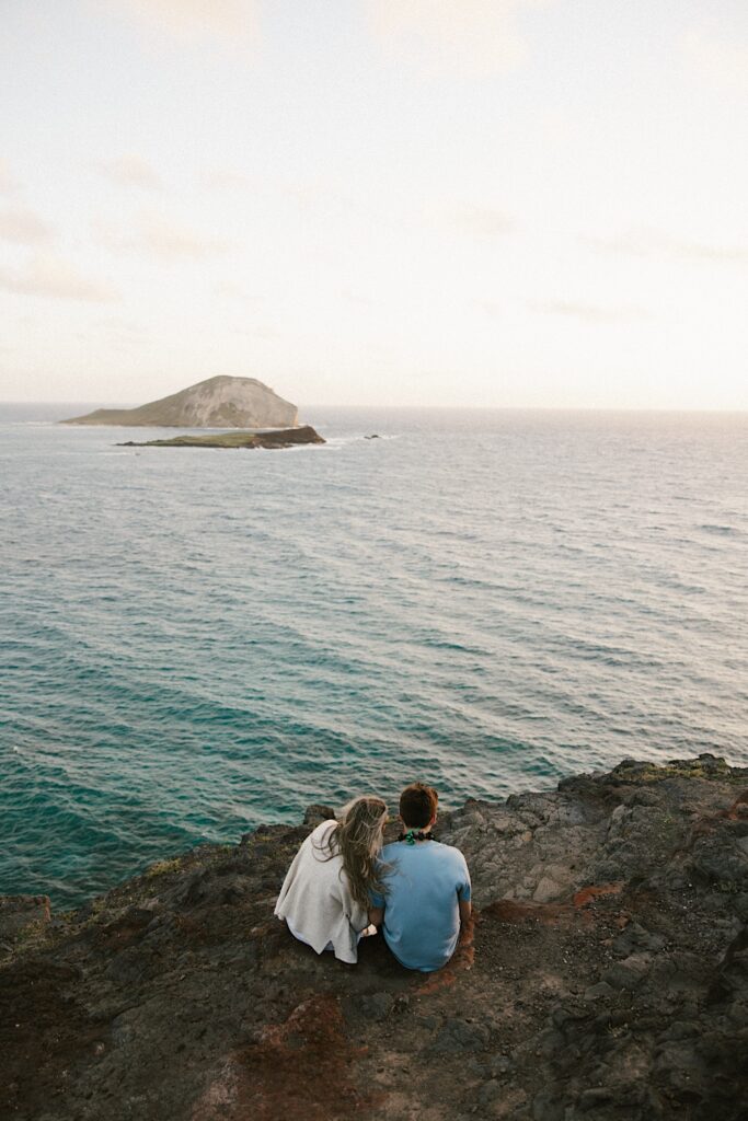 A couple sit next to one another atop a cliff in Hawaii and look out over the ocean and islands below