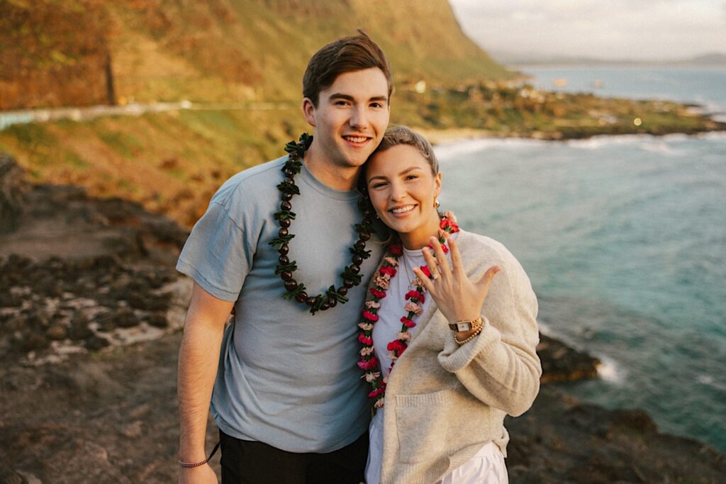 A couple embrace atop Makapuu Lookout after their proposal, the woman shows off her hand which has the engagement ring on it