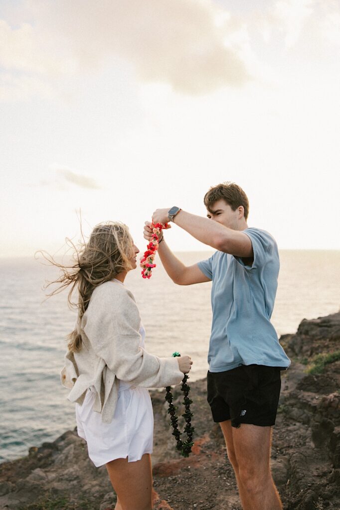 A couple holding flower leis smile at one another as they're about to put them on one another atop a cliff in Hawaii looking out over the ocean