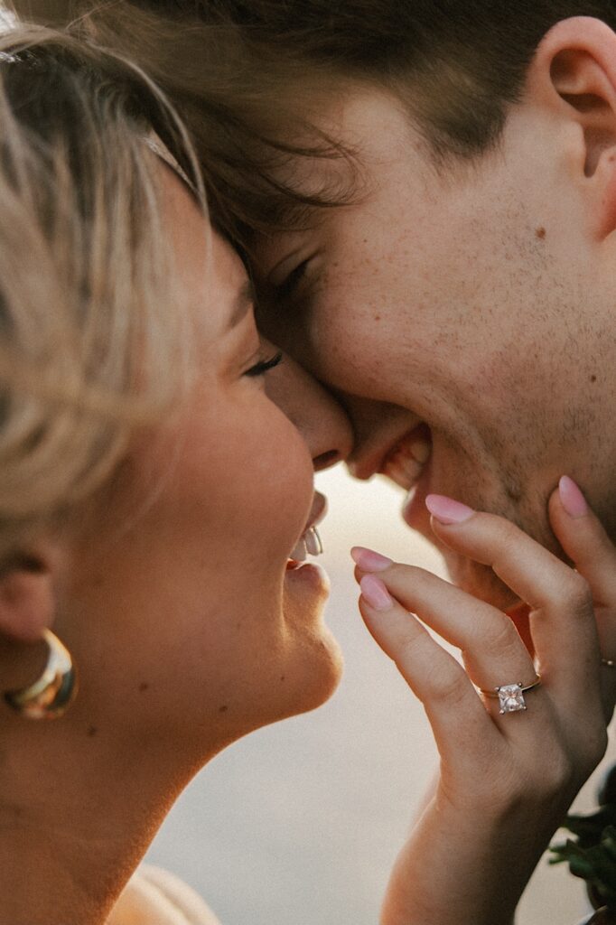 Close up photo of a couple smiling about to kiss one another, the woman shows off her hand which has an engagement ring on it