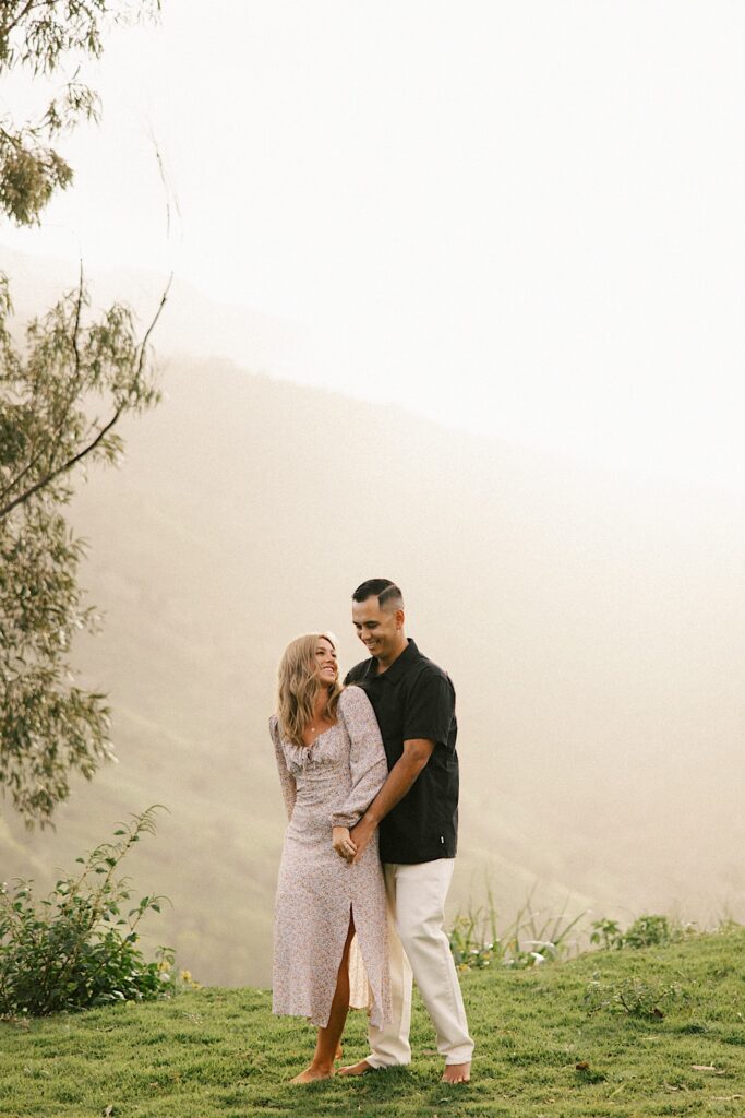 A couple smile as the man embraces the woman from behind, behind them are foggy mountains of Oahu