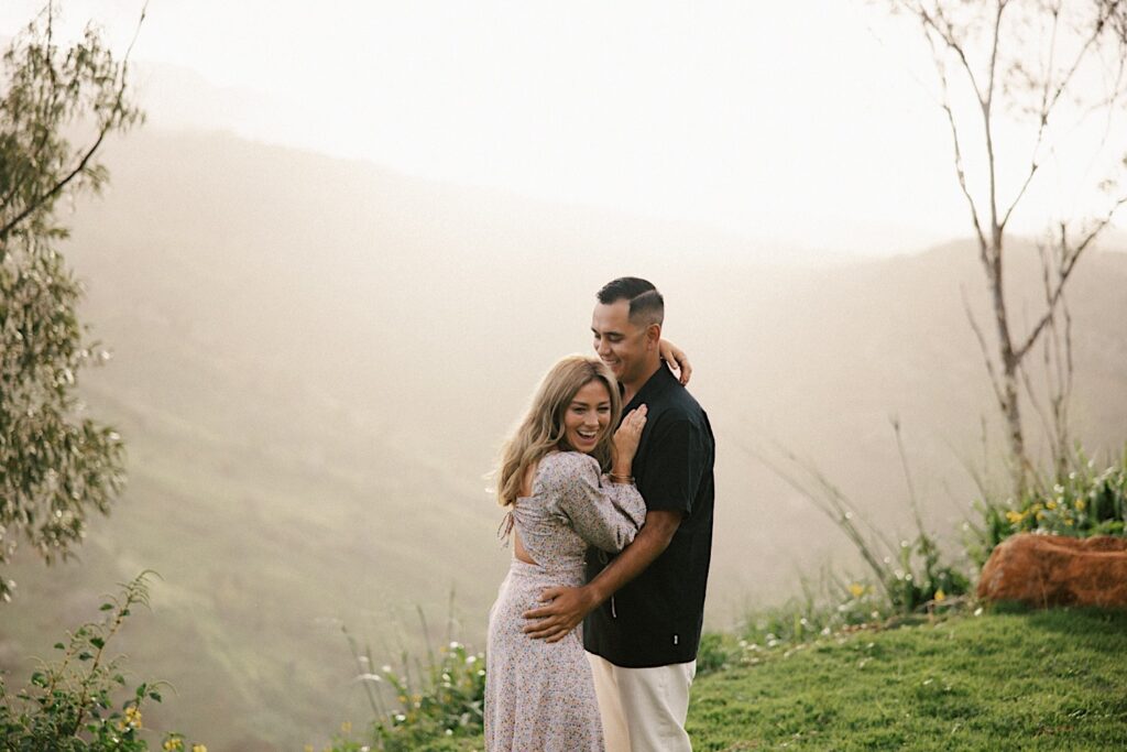 A couple embrace and laugh during their family session on Oahu, behind them are mountains covered in fog