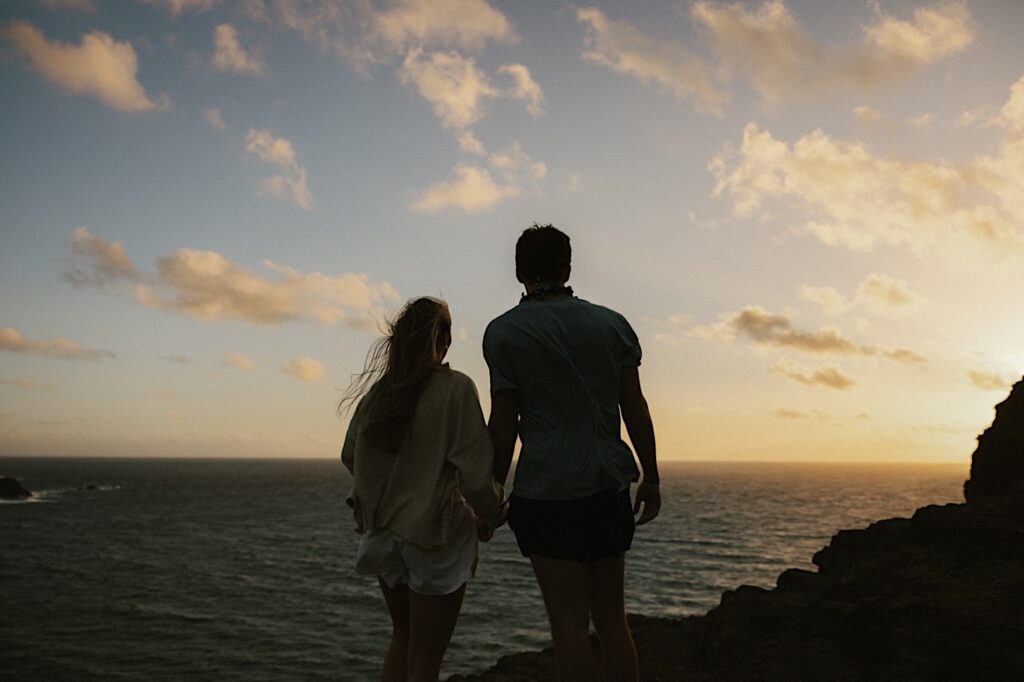 Silhouette of a couple on Makapuu Lookout holding hands and looking out at the ocean and sunset after their proposal