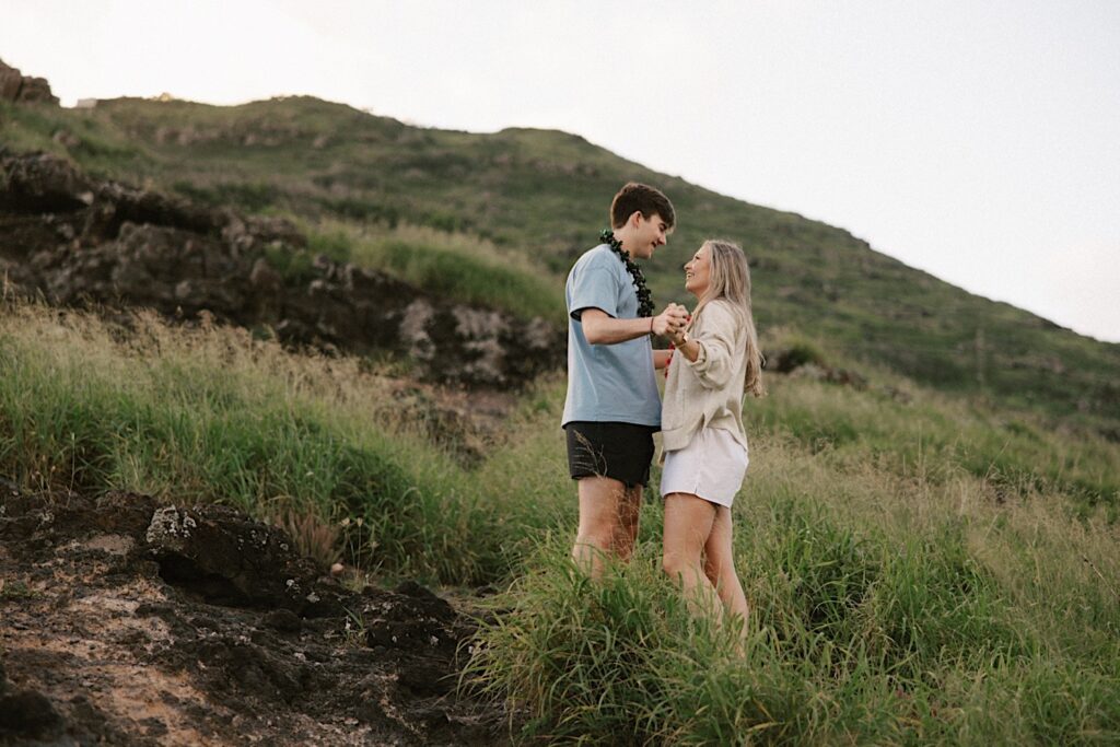 A couple in the grass on Makapuu Lookout smile and hold hands while looking at each other after their proposal
