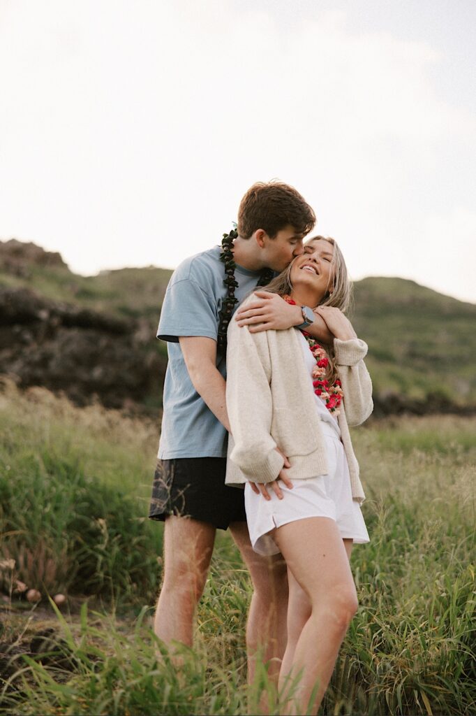 A couple embrace and the man kisses the girl on the cheek as she smiles while they stand in the grass on a Hawaiian Island