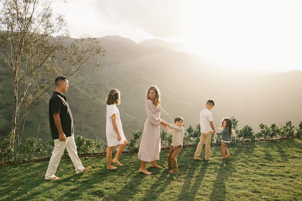 A family of six walk along a cliffside of Oahu that looks out over a mountain at sunrise during their family session