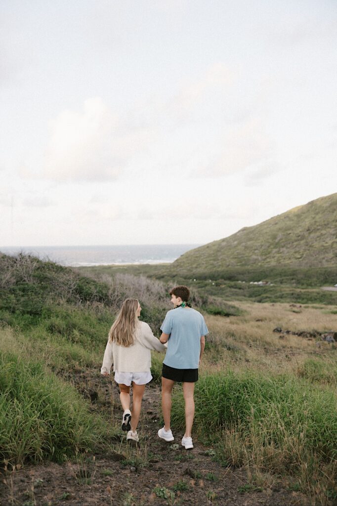 A couple walk down a grassy hill in Hawaii while holding hands and looking at one another