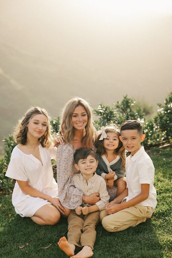 A woman sits on a cliff looking out over a mountain of Oahu surrounded by her 4 children who are all smiling at the camera