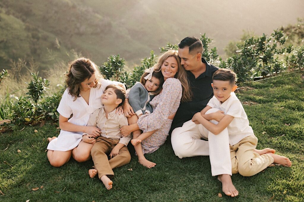 A family of 6 sit together and cuddle on a cliff looking out over a mountain of Oahu during their family session