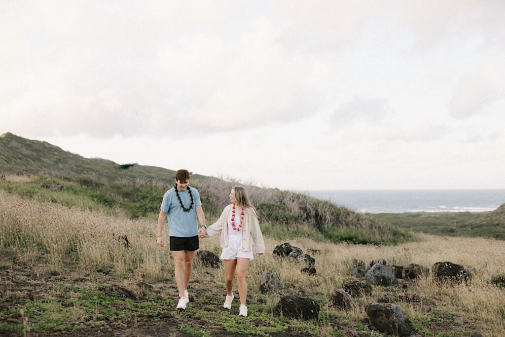 After their proposal on Makapuu Lookout in Hawaii a couple walk towards the camera and laugh while holding hands on a grassy path